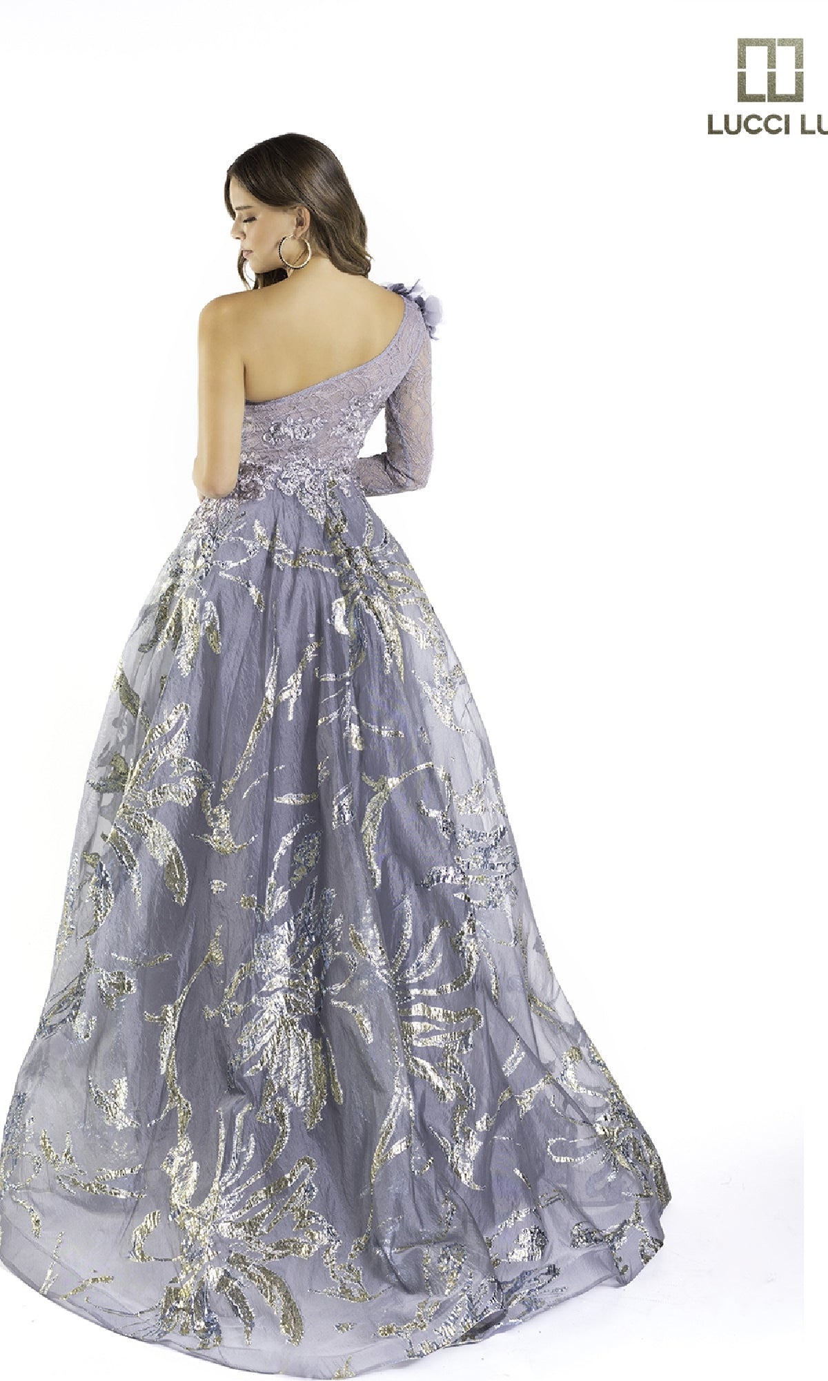 A woman stands facing away from the camera wearing the Lucci Lu C8054, a floor-length lavender lace prom dress with silver floral embroidery and a one-shoulder design.